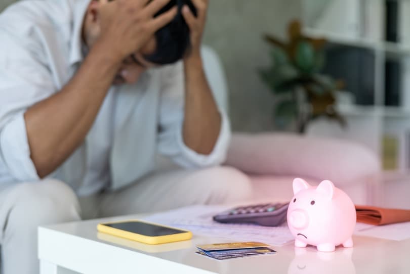 Muž sedí se sklopenou hlavou u stolu s pokladničkou, kartou a kalkulačkou, což znázorňuje finanční problémy a dluhy.