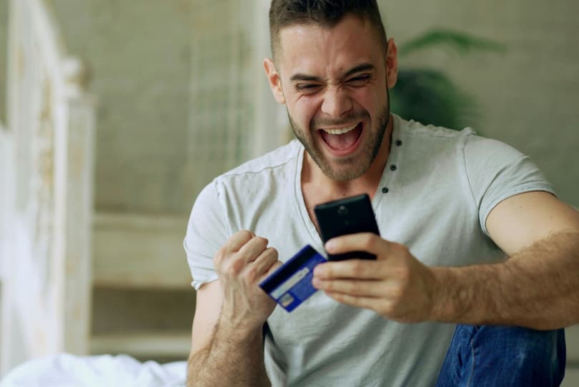 Muž s kreditní kartou v ruce a mobilním telefonem v druhé ruce, křičí radostí, pravděpodobně z úspěšného dokončení platby.