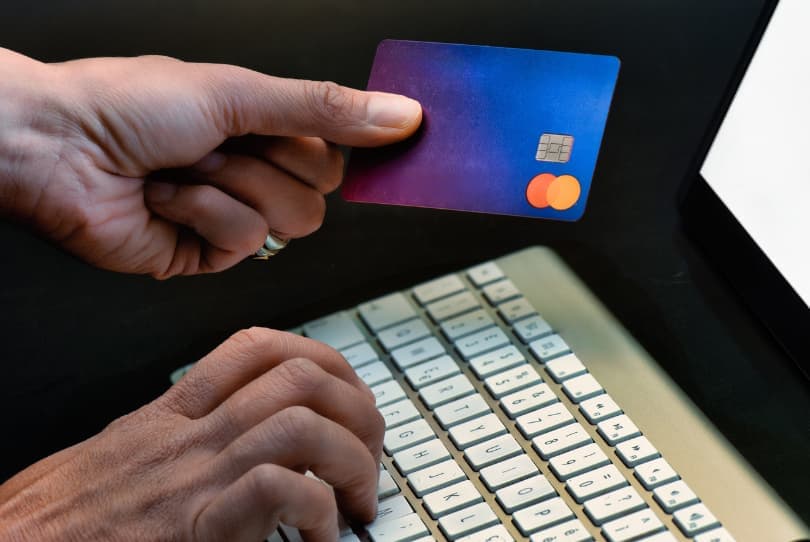 Ruka držící kreditní kartu, připravená na platbu online, přičemž na pozadí je vidět klávesnice počítače.